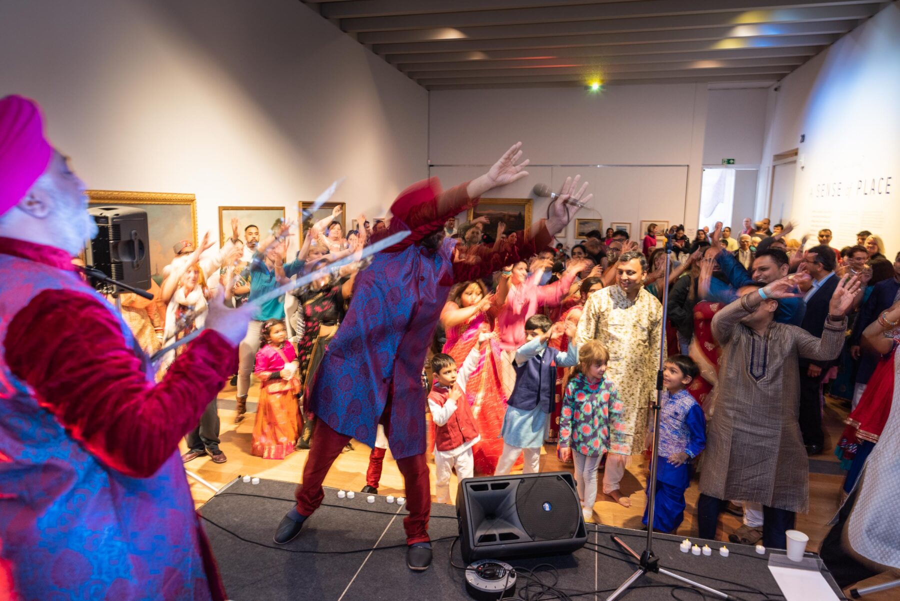 People smiling and laughing, dressed in Indian clothing dancing in a museum gallery, while a man in a turban leads the dance from a stage