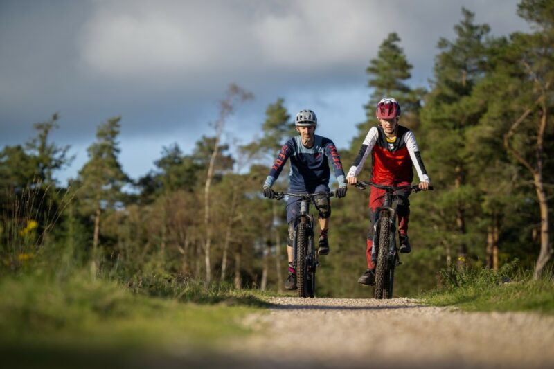 Two cyclist ride towards the camera on a wide flat forest trail