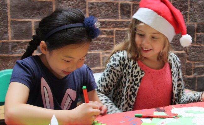 two young girls, one in a santa hat, making xmas crafts