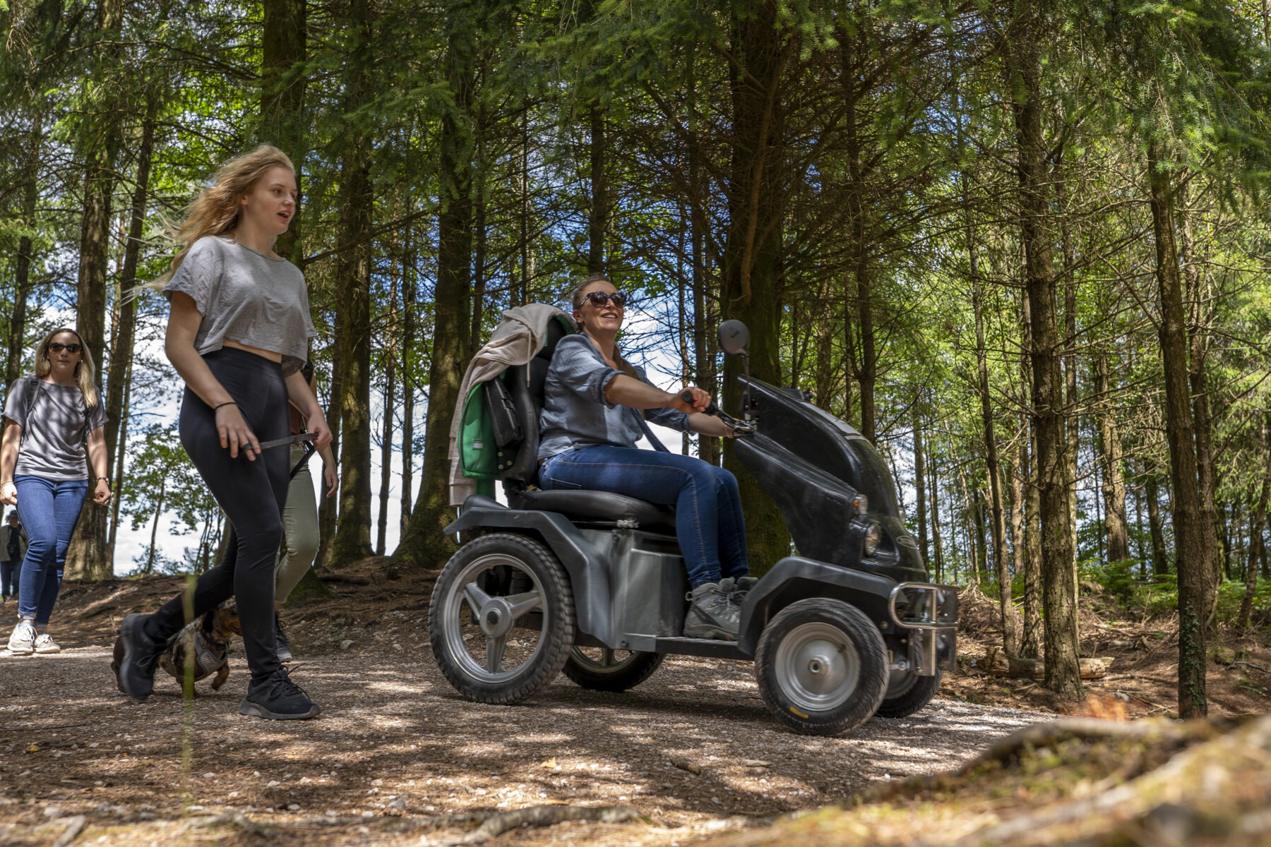 A young woman walks next to a woman on a mobility scooter in a conifer plantation