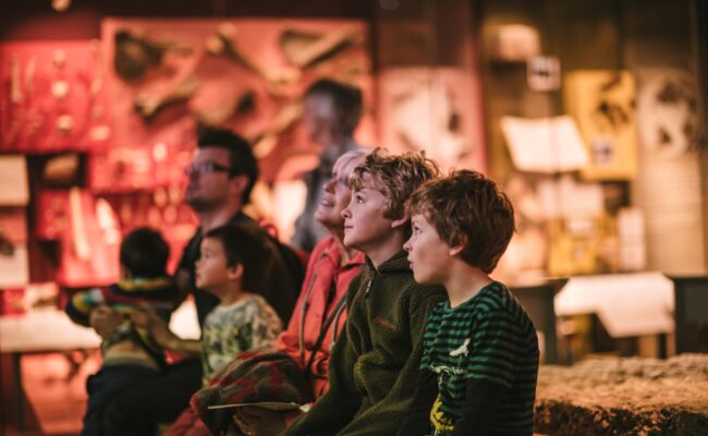 A family seated in a museum gallery, looking up at something. Behind them objects such as bones and fossils are visible behind glass
