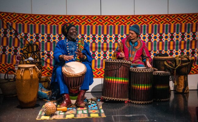 two men, one black and one white, seated in the museum courtyard playing djembe drums and laughing