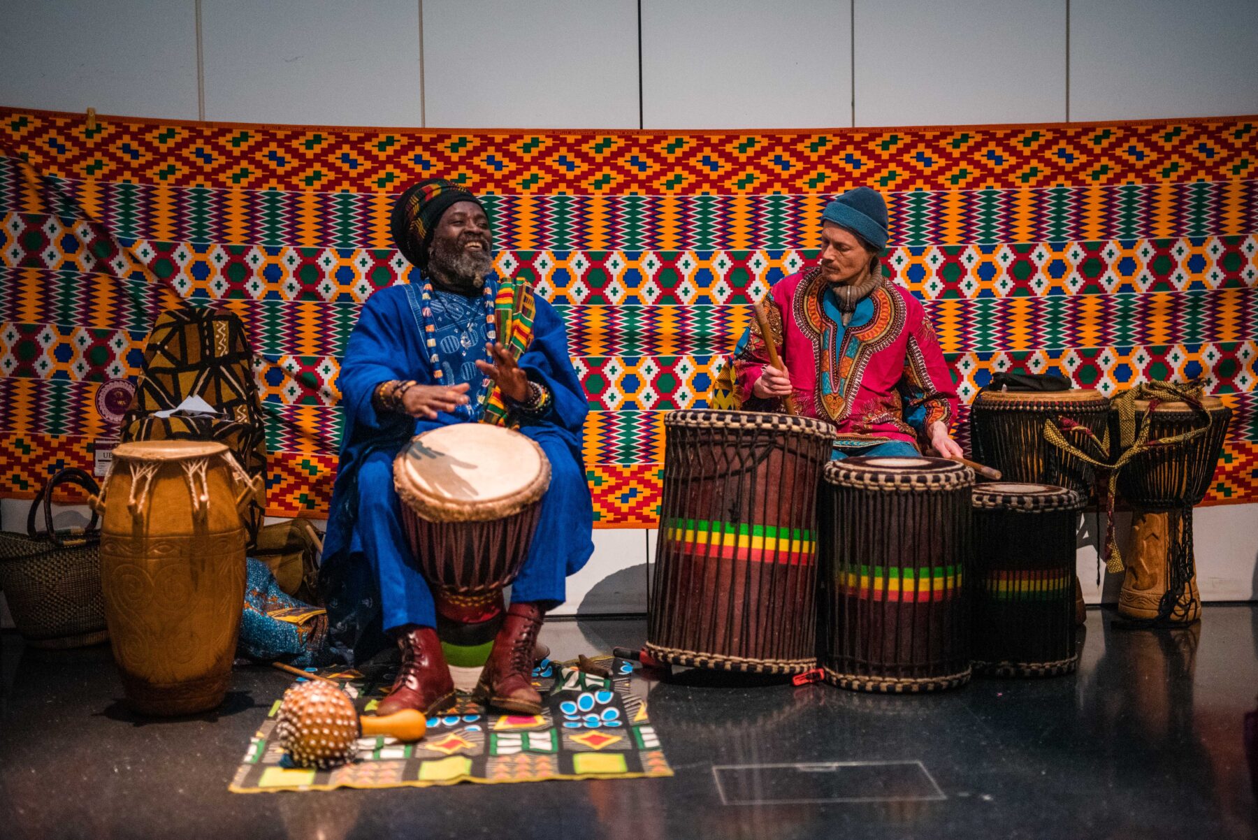 two men, one black and one white, seated in the museum courtyard playing djembe drums and laughing