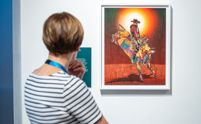 A woman in a striped top looks at an artwork featuring a cowboy as it hangs on a gallery wall