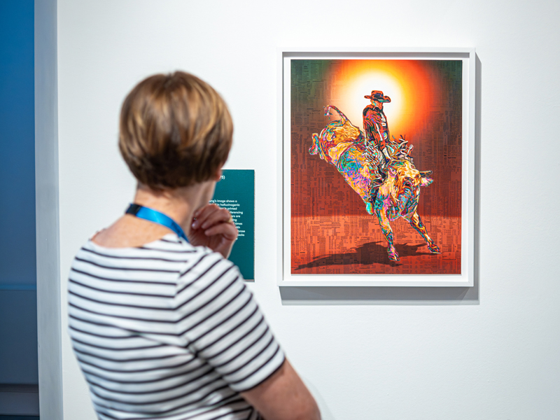 A woman in a striped top looks at an artwork featuring a cowboy as it hangs on a gallery wall
