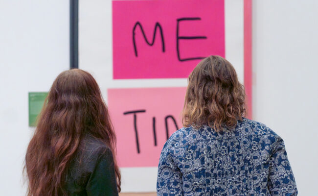 Two women look at a large artwork in pink and black featuring the words 'Me Time'