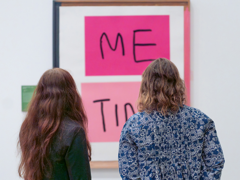 Two women look at a large artwork in pink and black featuring the words 'Me Time'