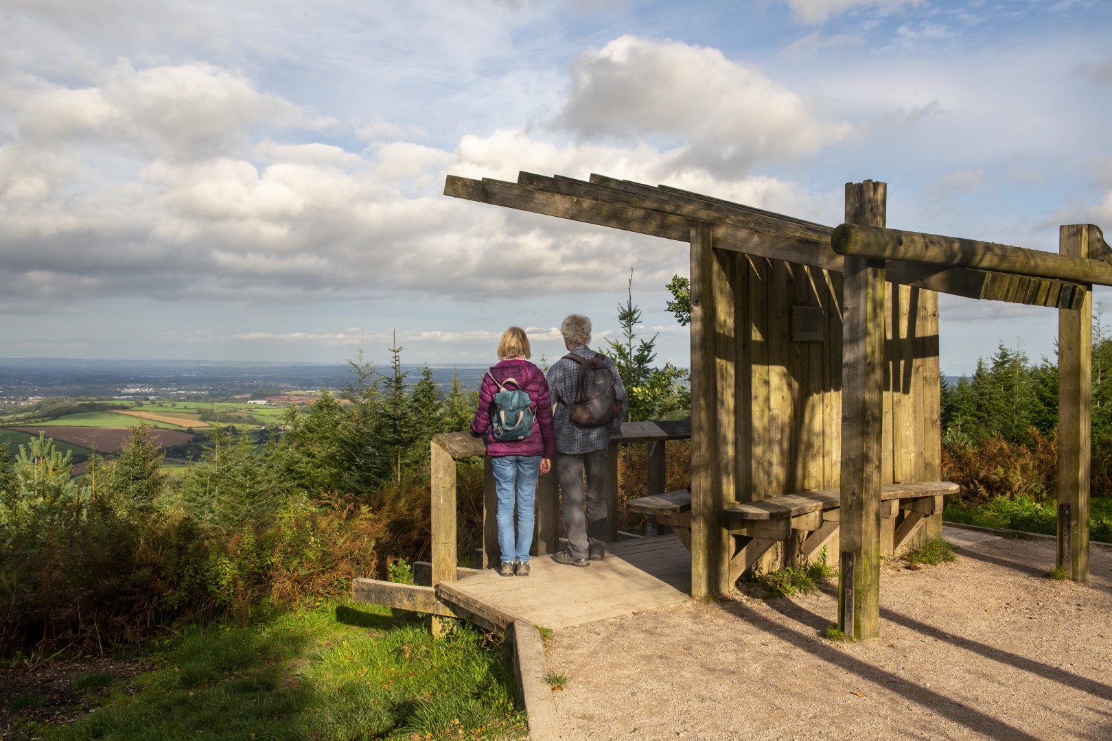 A couple stand under a wooden shelter looking at a far-reaching view