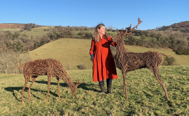Woman in a red dress stands next to two willow deer sculptures in Dartmoor