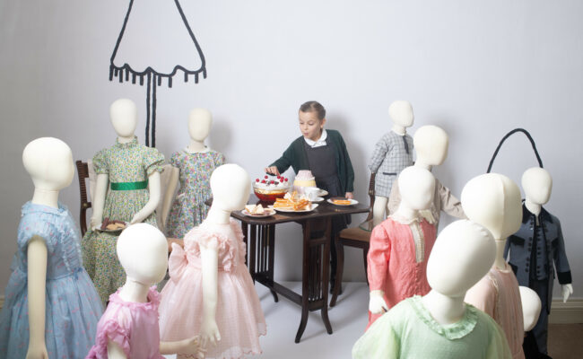 A girl picks at display food whilst surrounded by mannequins dressed in childrens clothing.