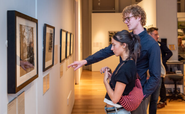 a young man and woman looking at framed art on a gallery wall; the man is pointing at the text below one of the artworks