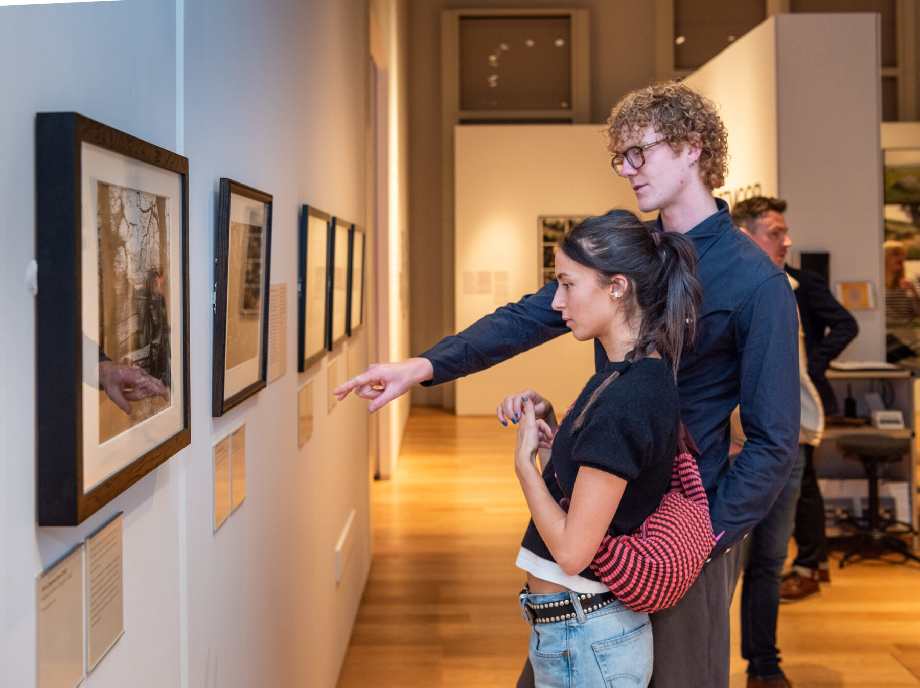 a young man and woman looking at framed art on a gallery wall; the man is pointing at the text below one of the artworks