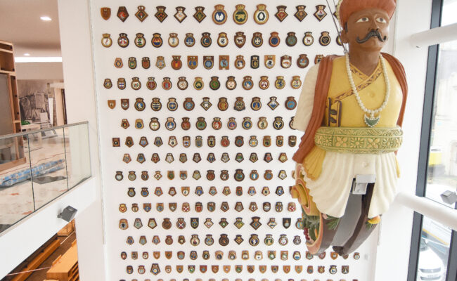 A large yellow, white and orange ship's figurehead hangs in front of a wall of ship's badges