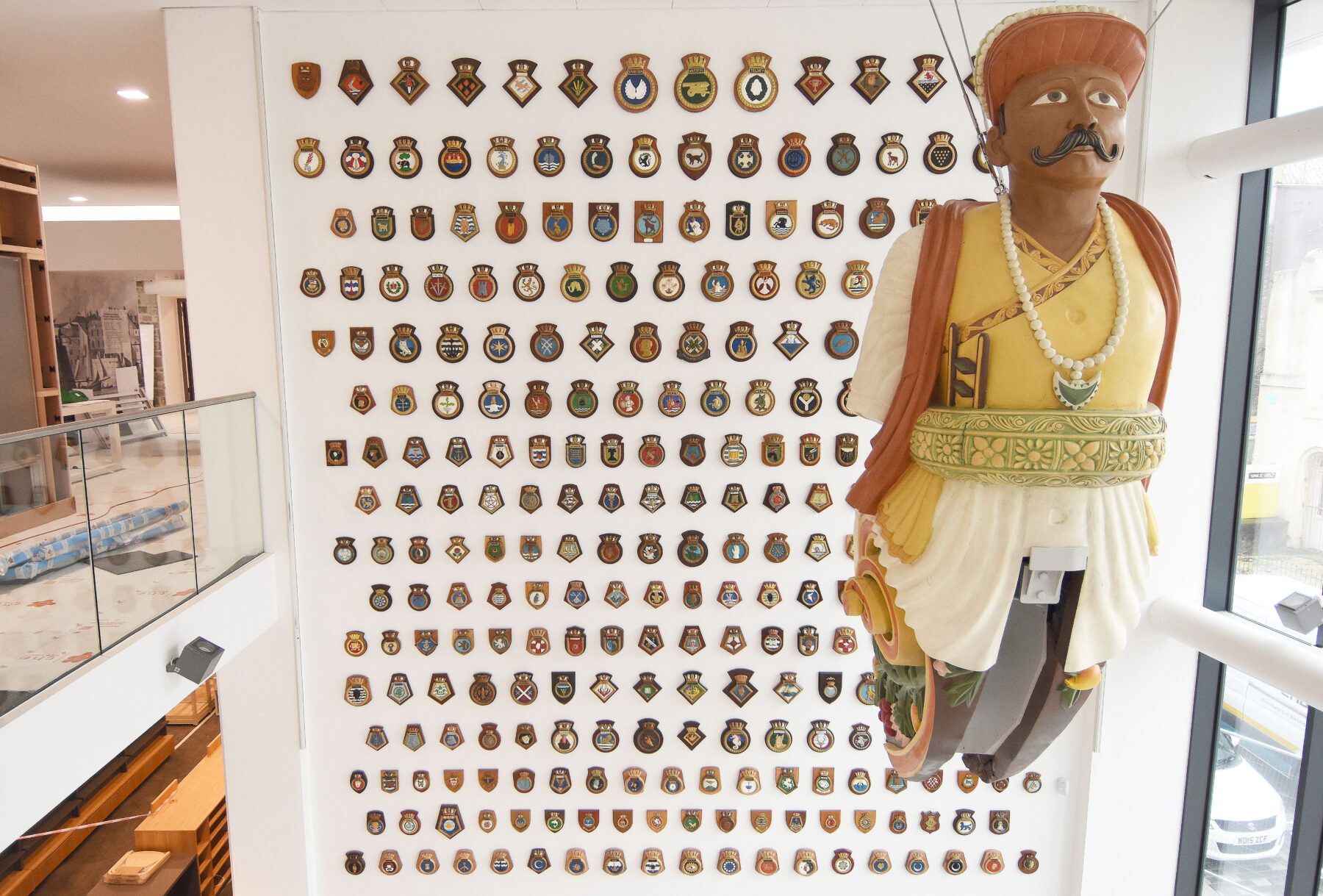 A large yellow, white and orange ship's figurehead hangs in front of a wall of ship's badges