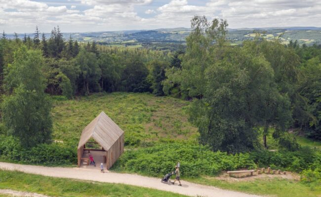 A timber shelter viewed from the air as a man pushes a pushchair along a trail