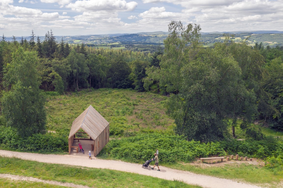 A timber shelter viewed from the air as a man pushes a pushchair along a trail
