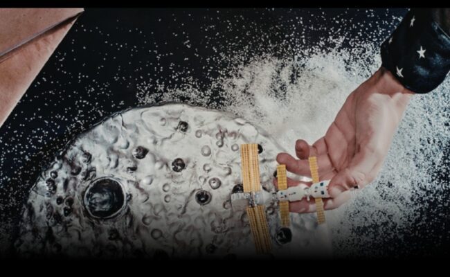 a child's hand holding a cardboard space craft above a cardboard moon