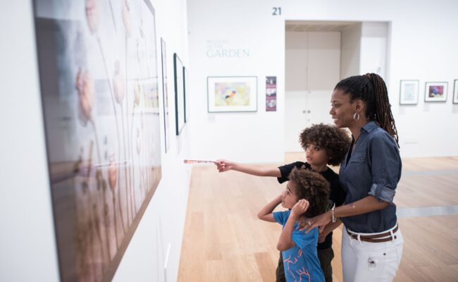 a woman and two boys in a gallery, pointing at a large artwork on the wall